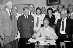 black and white image of Roosevelt signing documents at table with large group of men and one woman in background